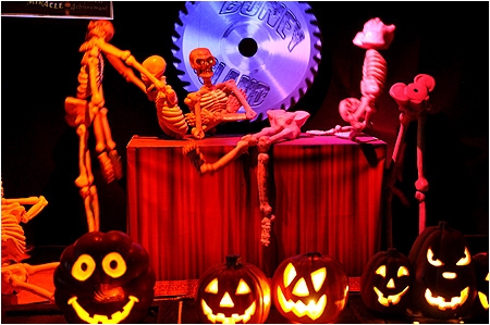 Skeletons doing their best to work a big circular saw while some pumpkins smile on in the foreground.